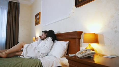 young man and woman rest in a hotel. lovely couple in white dressing gowns lying on bed and talking. happy newlyweds spend time together during honeymoon.