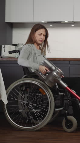 female person with disability vacuums in kitchen sitting in wheelchair. young housewife cleans apartment carefully using modern equipment side view