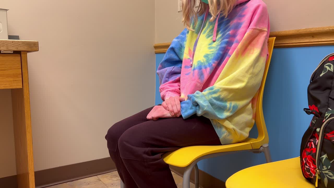 Young kid child girl or boy in covid-19 mask at Doctor’s office hospital visit waiting on yellow chair nervously making fists and shifting in seat.
