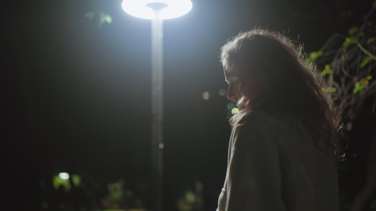 Woman walking through dim park at night with streetlight casting glow around, highlighting her silhouette against darkness, with faint figures in blurred background