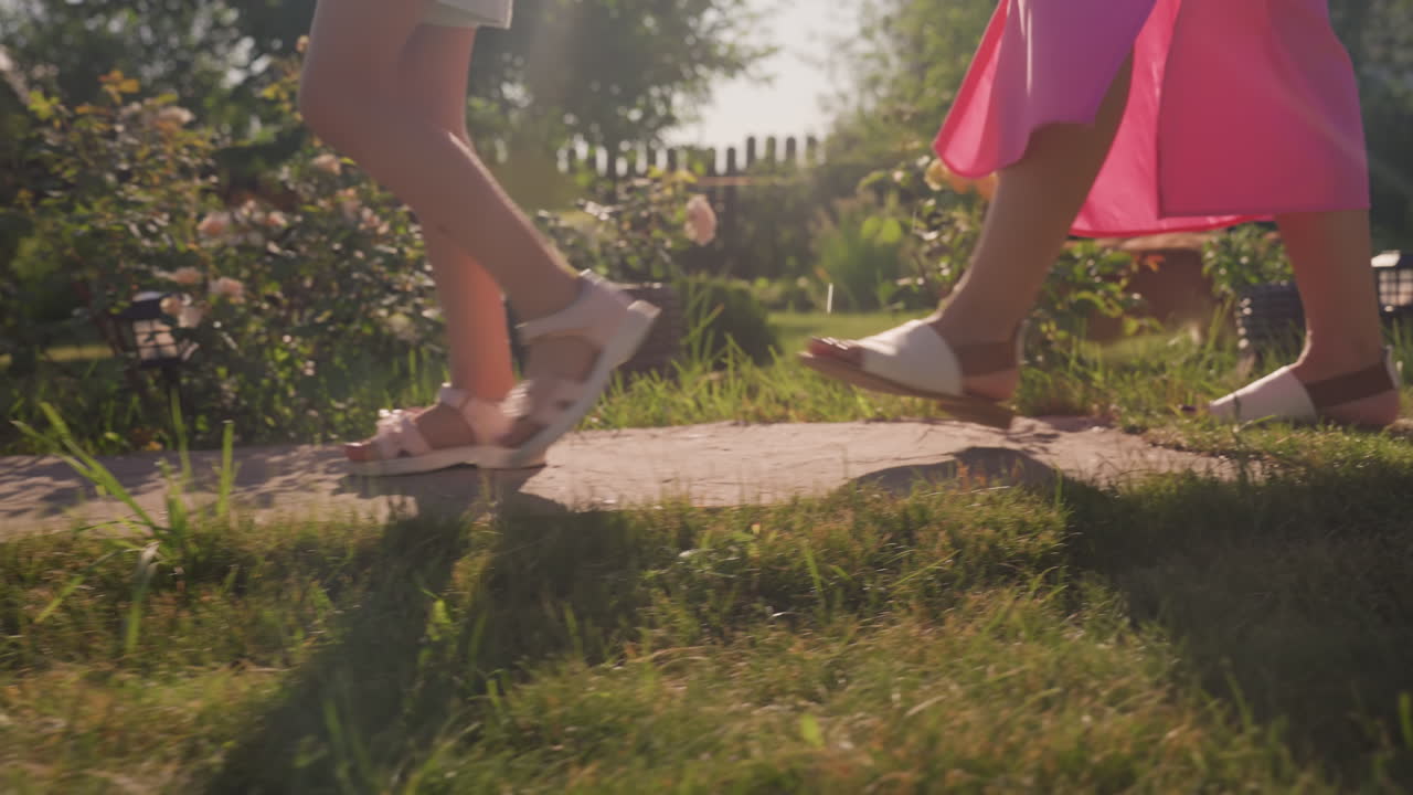 Little Caucasian Girl Walking With Corgi On Stone Path In Sunlit Garden, Close Shots Of Sandals And Small Dog Following Step By Step, Warm Summer Light, Green Grass, Fence And Floral Beds