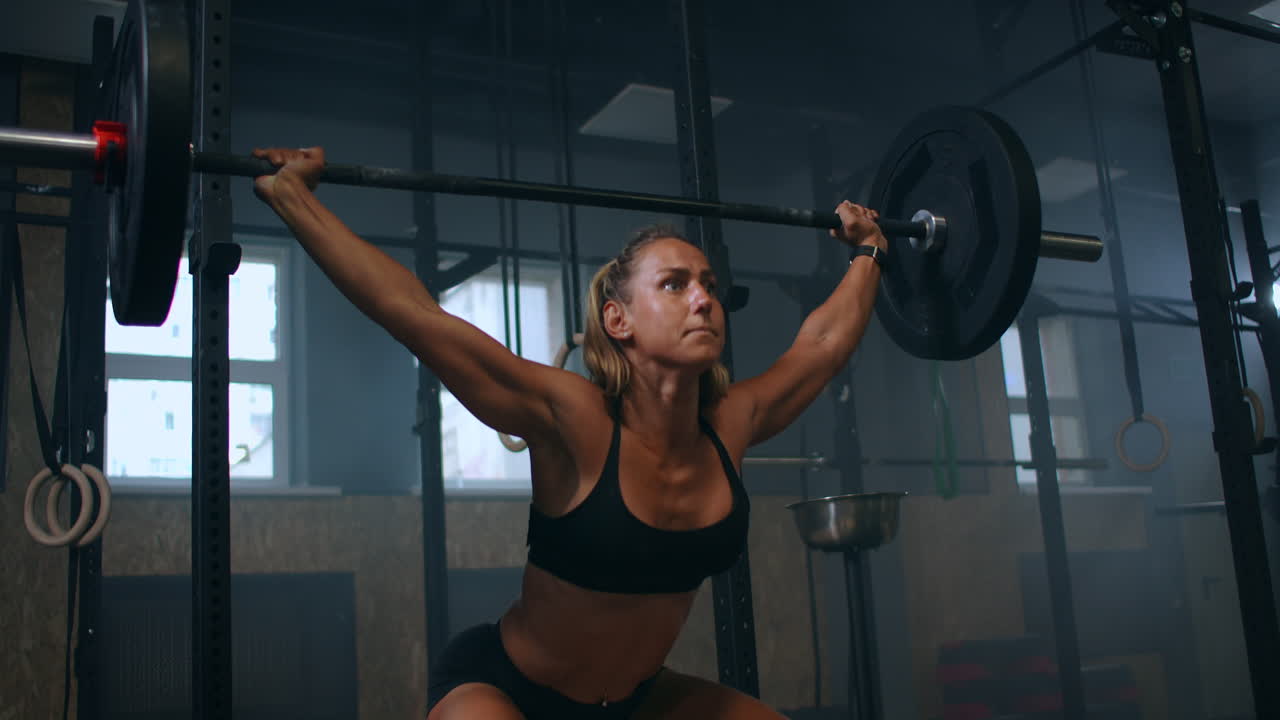 Ralenti : entraînement de squats avec barre au-dessus de la tête. Femme musclée soulevant une barre au-dessus de la tête pour entraîner ses épaules à la salle de sport. Femme de fitness confiante pratiquant un entraînement de force avec équipement lourd.
