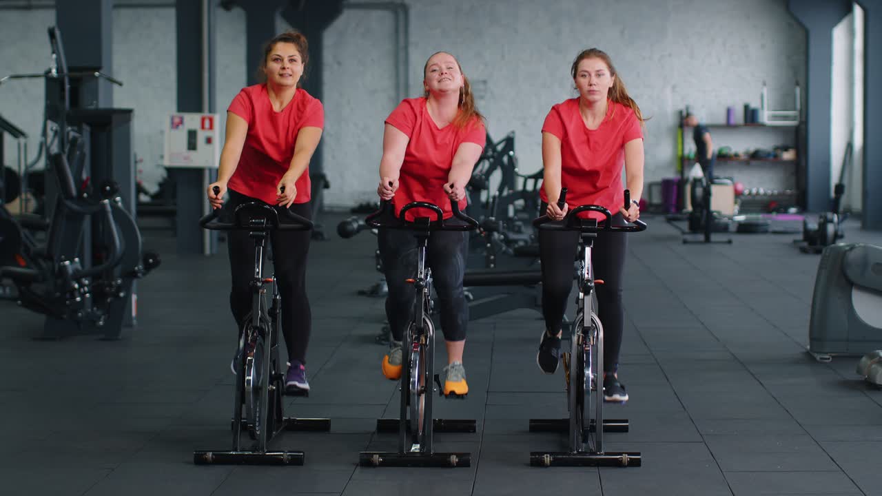 건강한 백인 여성 그룹이 체육관에서 정지 자전거 기계 자전거에서 운동을합니다.