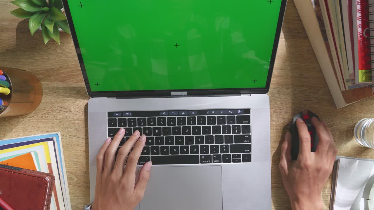 Top down view of a laptop computer with mock up green screen chromakey display on a wooden office desk next to notebook with pens, glasses, and a glass of water. Slow zoom out, close up