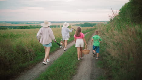 back view of family walking home along a dirt path through lush countryside, two women in hats walk with a dog on a leash, while a young girl carries a basket and a boy plays with a stick