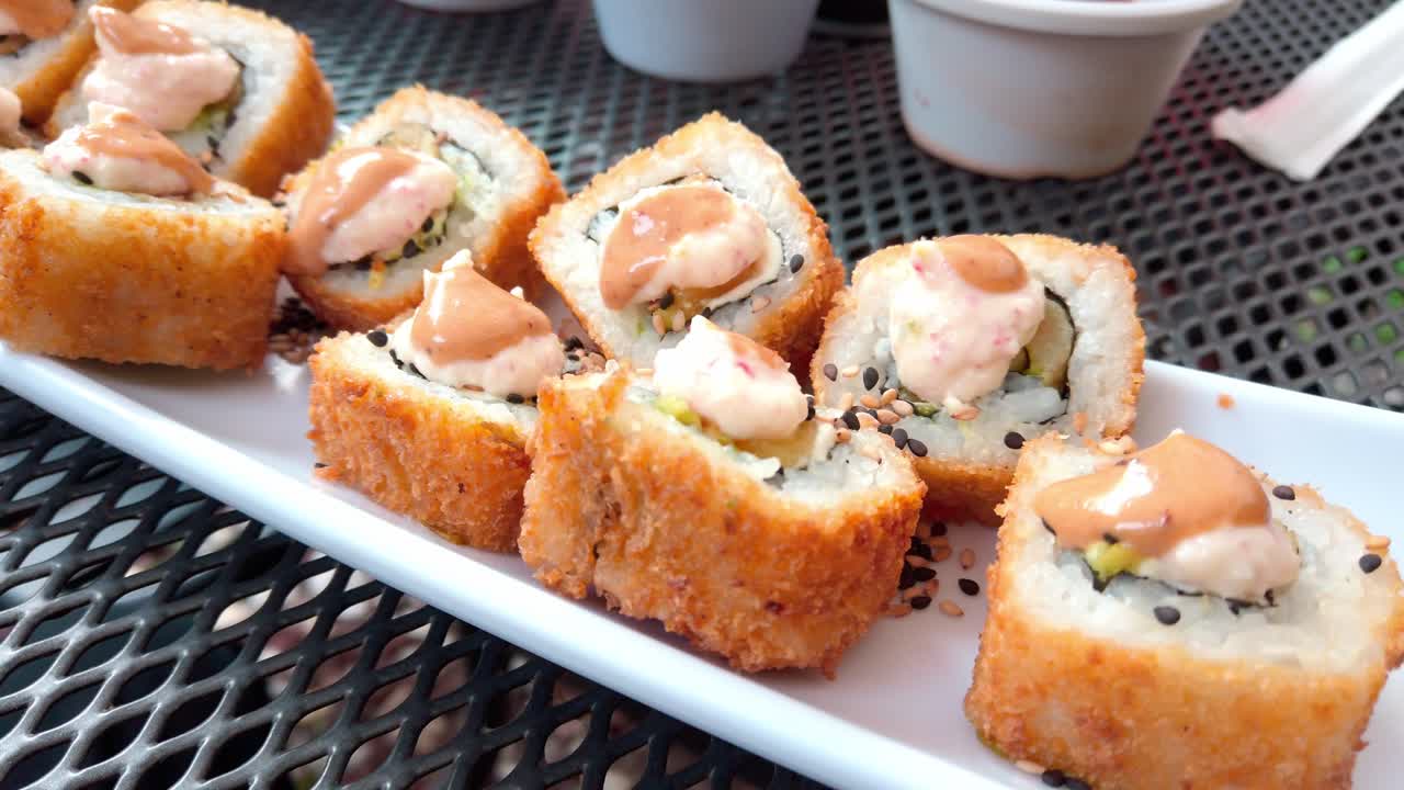 Appetizing close-up of fried sushi rolls topped with creamy sauce, served on a white plate over a metal mesh table.