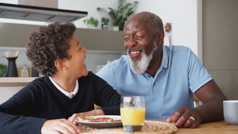 Portrait-Of-Smiling-Grandfather-Sitting-In-Kitchen-With-Grandson-Eating-Breakfast-Before-School