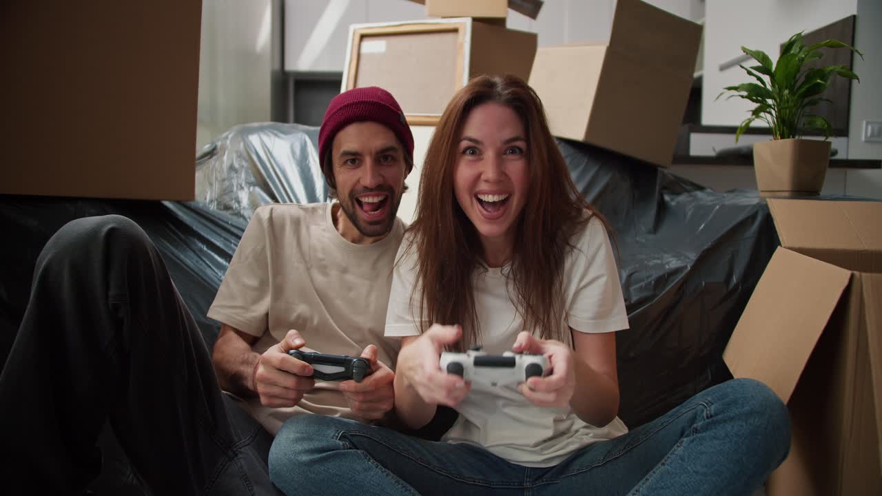 A happy brunette girl in a white T-shirt plays with her boyfriend with stubble in a video game using two joysticks near them there are a large number of boxes and a sofa in a black cover after moving