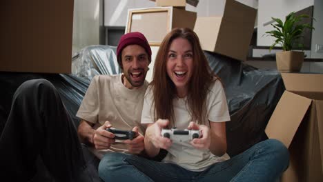 A-happy-brunette-girl-in-a-white-T-shirt-plays-with-her-boyfriend-with-stubble-in-a-video-game-using-two-joysticks-near-them-there-are-a-large-number-of-boxes-and-a-sofa-in-a-black-cover-after-moving