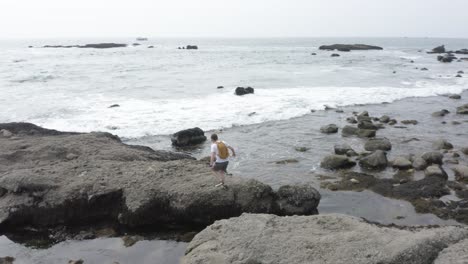 Ein-Junger-Weißer-Mann,-Der-Mit-Einem-Gelben-Rucksack-Und-Einem-Drohnencontroller,-Der-Mit-Brechenden-Wellen-An-Seinem-Hals-Hängt,-Am-Meer-Entlang-Wandert