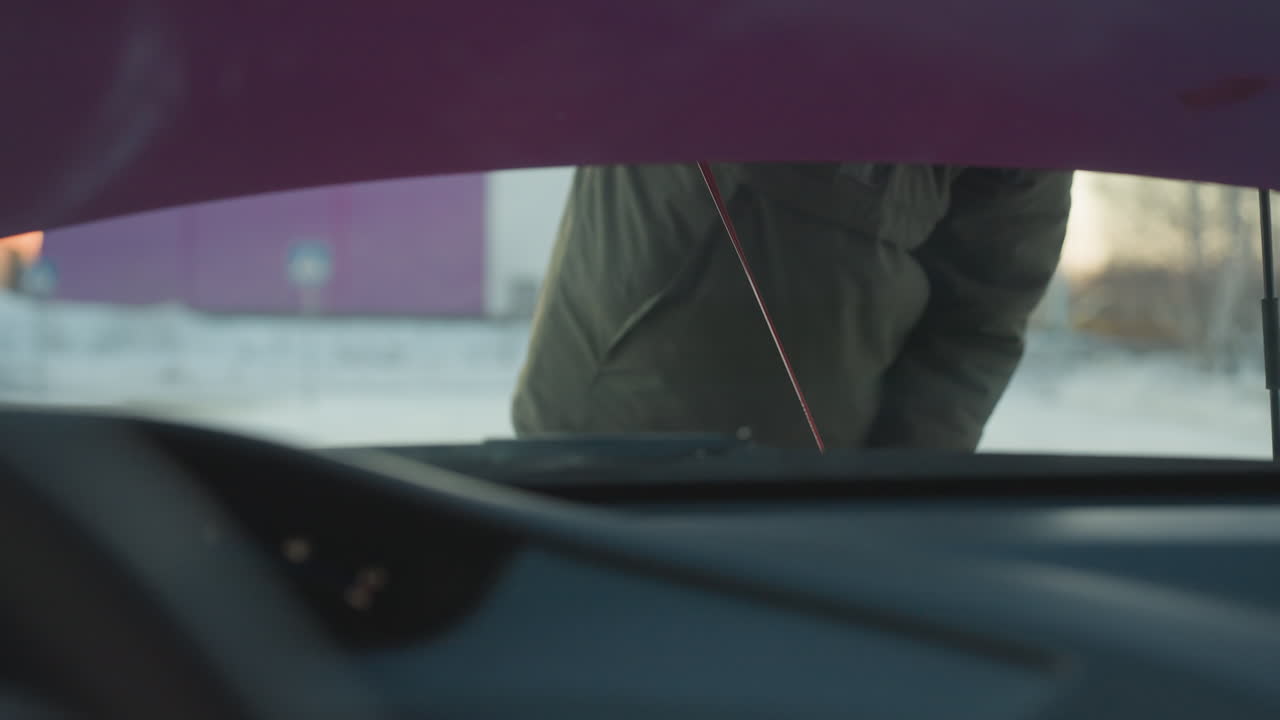 Close up of person removing rubber oil gauge from car engine bay to check oil level during cold winter maintenance outdoors with snow in background and raised bonnet visible in soft light
