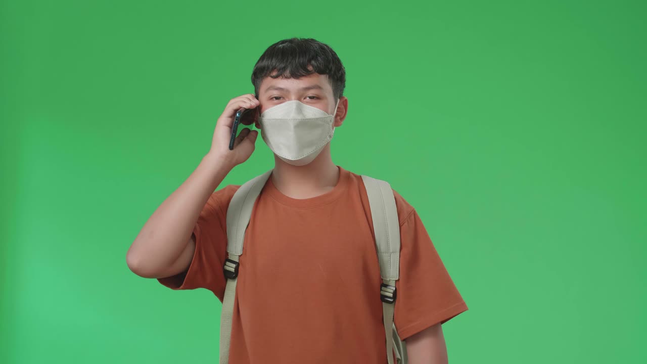 The Front View Of Asian Boy Student Wearing A Mask, Backpack And Brown T-Shirt Talking On Mobile Phone While Walking To School On Green Screen Chroma Key.