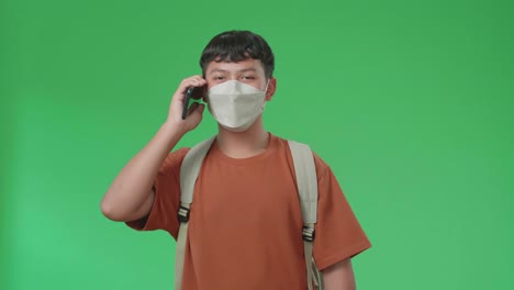 the front view of asian boy student wearing a mask, backpack and brown t-shirt talking on mobile phone while walking to school on green screen chroma key.