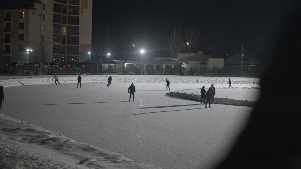 Blurred silhouette in foreground watches group of skaters glide on snow covered outdoor ice rink illuminated by overhead floodlights against dark urban night sky