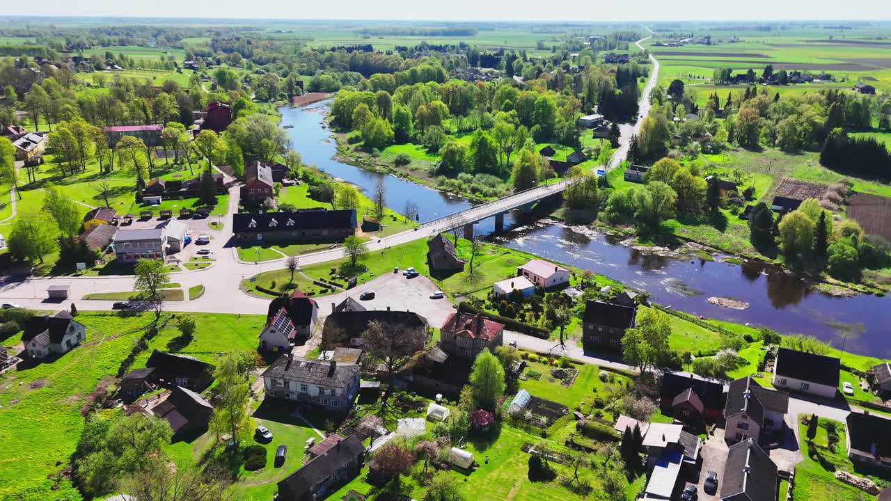 A Peaceful River Winds Beneath a Connecting Bridge, Framed by Quaint Houses and Verdant Scenery in Biržai, Lithuania - Orbit Drone Shot