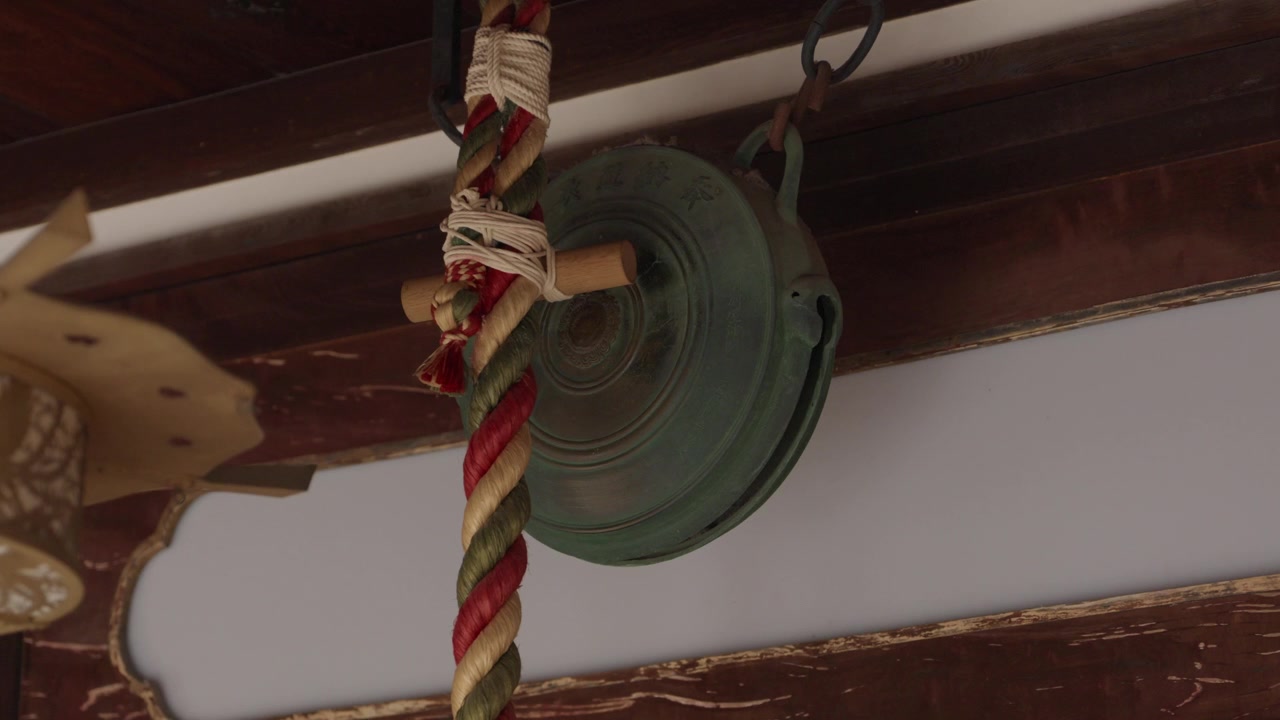 Campana votiva in bronzo patinato con una corda intrecciata colorata appesa nella sala delle offerte di un santuario giapponese.
