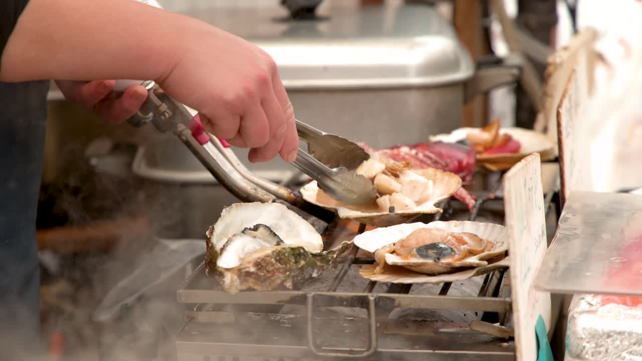 A market vendor uses a blowtorch and tongs to grill fresh shellfish on a grill
