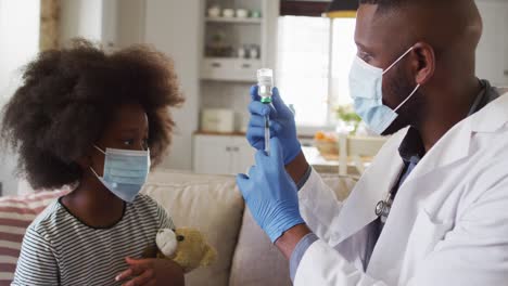 Afroamerikanischer-Arzt-Mit-Gesichtsmaske,-Der-Zu-Hause-Die-Spritze-Mit-Covid-19-Impfstoff-Füllt