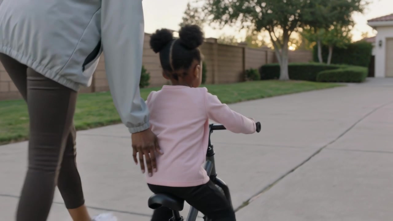 Guiding adult steadying child pedaling black balance bike on driveway, teaching balance, jacket