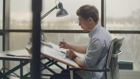 architect-man-Standing-working-with-blueprints-sketching-a-construction-project-on-wood-desk-at-home-office.Construction-design-concept.vintage-color-tone.-Engineer-works-in-a-bright-office-with-a-large-window-concentrates-and-draws-blueprints.-Workplace-of-an-architect-or-designer:-loft-style-minim