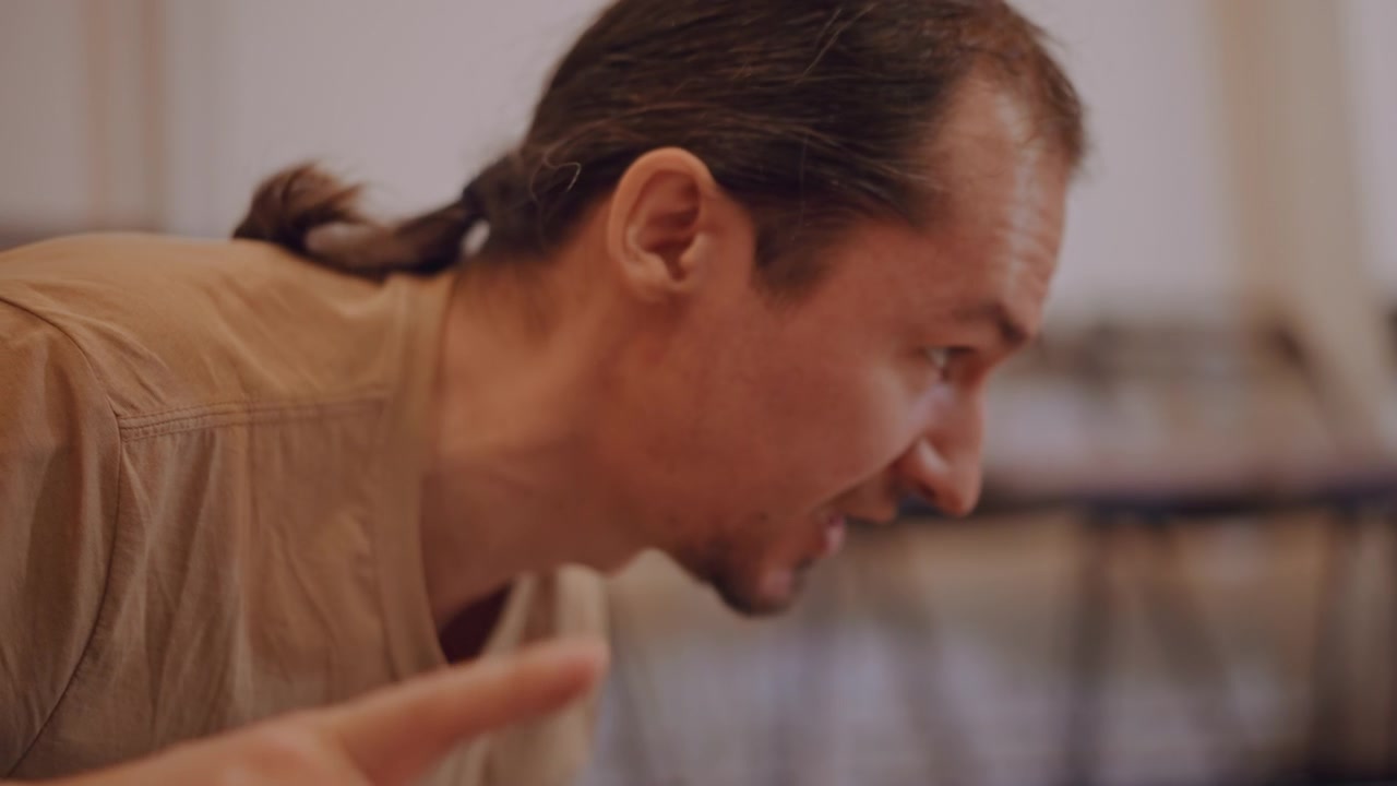 Caucasian music coach leaning forward, pointing and instructing in small rehearsal classroom, intense expression, ponytail, casual shirt, blurred chairs in background, demonstrating rhythm and posture