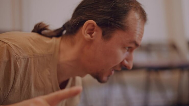 Caucasian music coach leaning forward, pointing and instructing in small rehearsal classroom, intense expression, ponytail, casual shirt, blurred chairs in background, demonstrating rhythm and posture