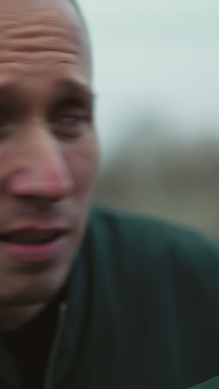 una vista cercana de un hombre apuntando una pistola con intenso enfoque y determinación, el entorno al aire libre incluye un fondo borroso de una valla y un paisaje tranquilo