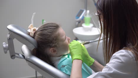 Female-dentist-drilling-tooth-of-cute-little-girl-at-the-dentist-cabinet-and-uses-saliva-ejector.-Shot-in-4k