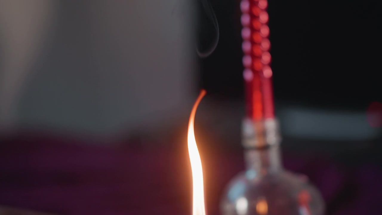 Primer plano de la llama de una vela cerca de una botella de vidrio, cuentas rojas sobre terciopelo, parpadeo lento con humo fino, brasa rizándose de la mecha, atmósfera cinematográfica y de ambiente para la lectura de la fortuna y el ritual de adivinación.