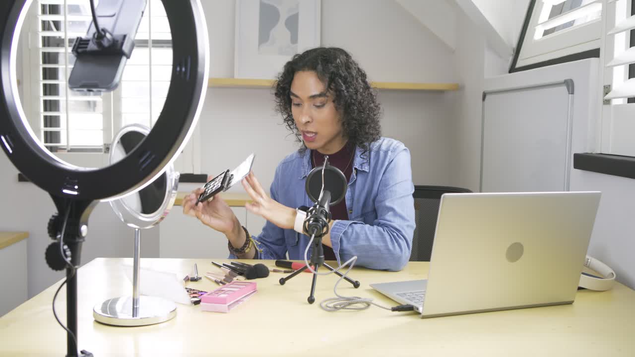Non-binary creator picking up palette, rotating shades, opening red-lid tube at desk with mic