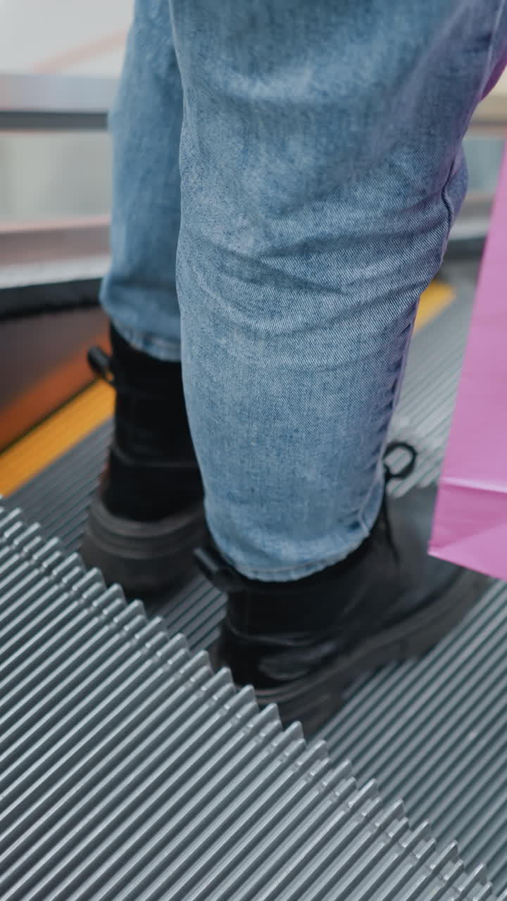 Close-up of lower legs in black boots and jeans, carrying a pink shopping bag while gently dangling it on a moving escalator in a bustling mall