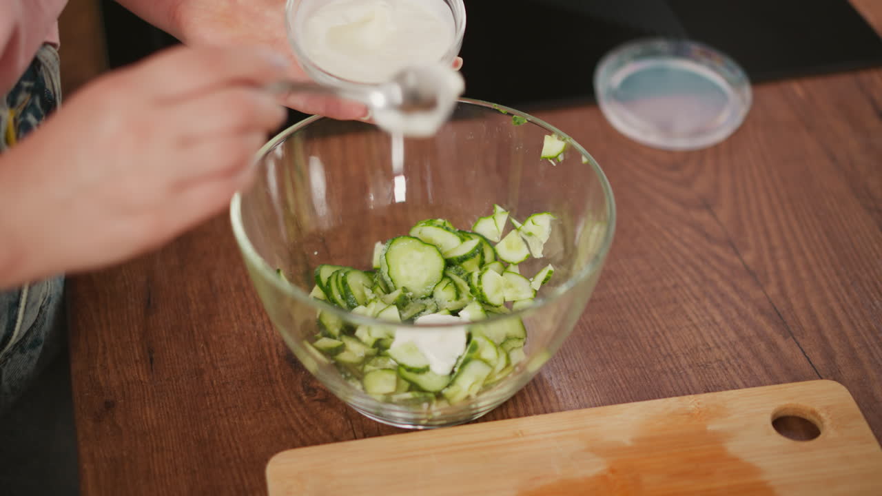 partial view of person holding spoon with creamy white dressing above glass bowl filled with sliced cucumber on wooden table, preparing salad with fresh ingredients in kitchen scene