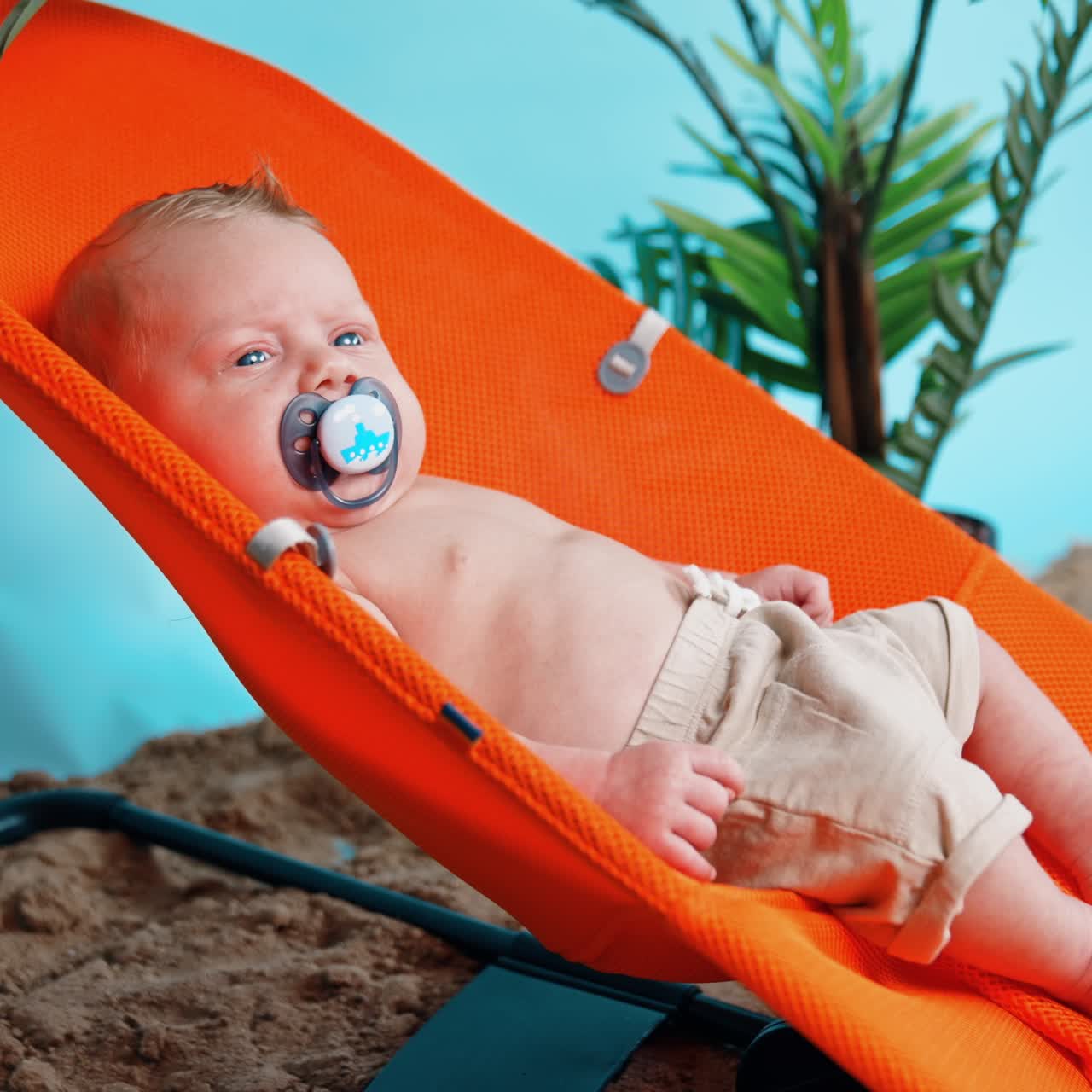 Lovely baby boy with pacifier in mouth lies in chair. Infant is winking from the bright light