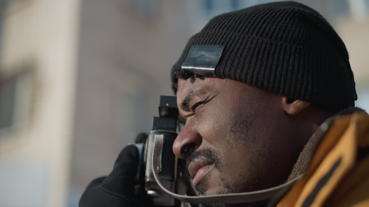 side view of man holding camera lens with gloved hands, eyes fixed on viewfinder under sunny sky, wearing winter coat and beanie capturing shot on snowy street with blurred cars in background