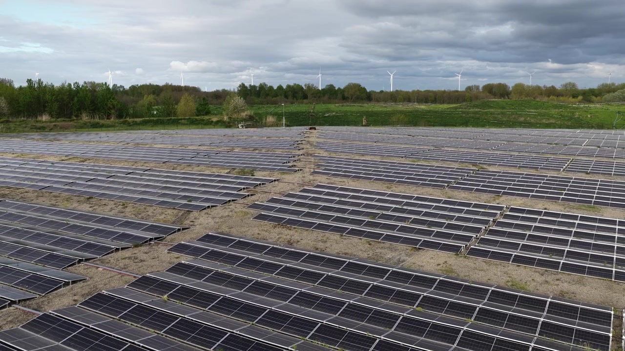En stor solcellepark med fotovoltaiske paneler og vindmøller i horisonten under en overskyet himmel, der illustrerer storstilet produktion af vedvarende energi og bæredygtig energiteknologi.