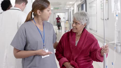 happy diverse female nurse talking to senior patient with drip and walking in slow motion, unaltered