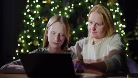 mom and daughter are looking at the laptop screen together. sitting in front of the blurred lights of the christmas tree