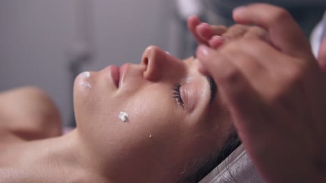 close up view of a woman with her eyes closed lying on the couch while professional cosmetologist is applying face cream and