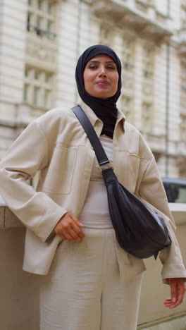 Vertical-Video-Portrait-Of-Smiling-Muslim-Businesswoman-Wearing-Hijab-And-Modern-Business-Suit-Standing-And-Putting-Hand-In-Pocket-Outside-City-Office-Buildings