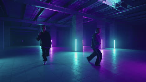 two hip-hop dancers in neon light dance together in blue and purple colors. women enjoying hip hop moves in dark studio with smoke and lighting.