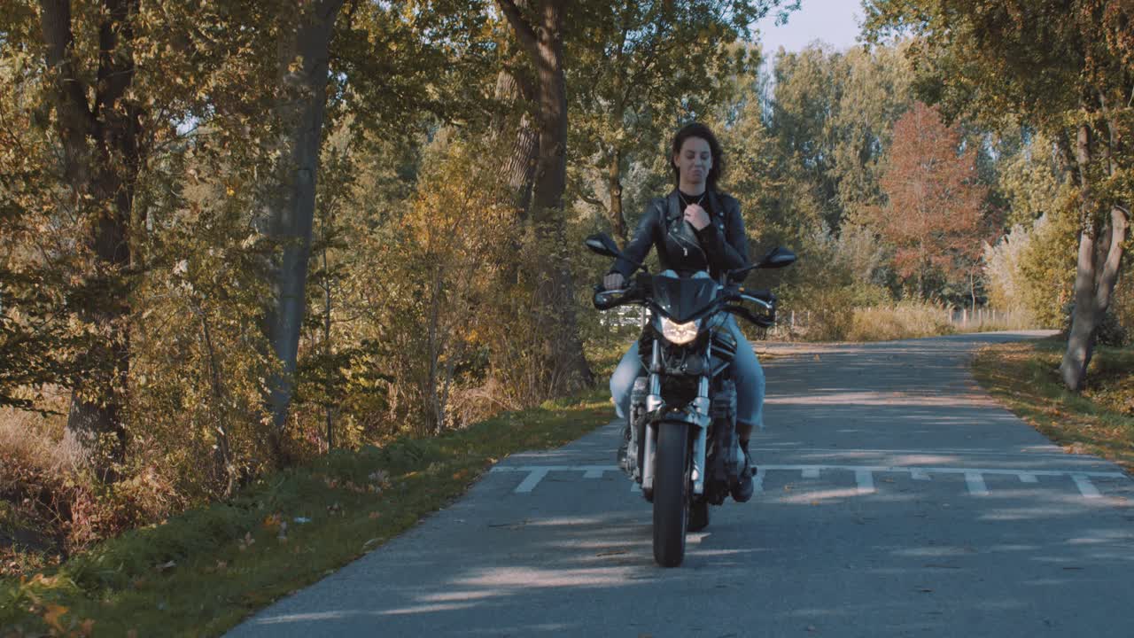 mujer joven europea muy sonriente conduciendo una moto con chaqueta de cuero en el bosque con hojas de otoño doradas vibrantes y coloridas en el día soleado-6