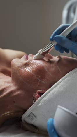 the esthetician is applying a clear treatment gel to the marked areas of the client's face using a brush - close up