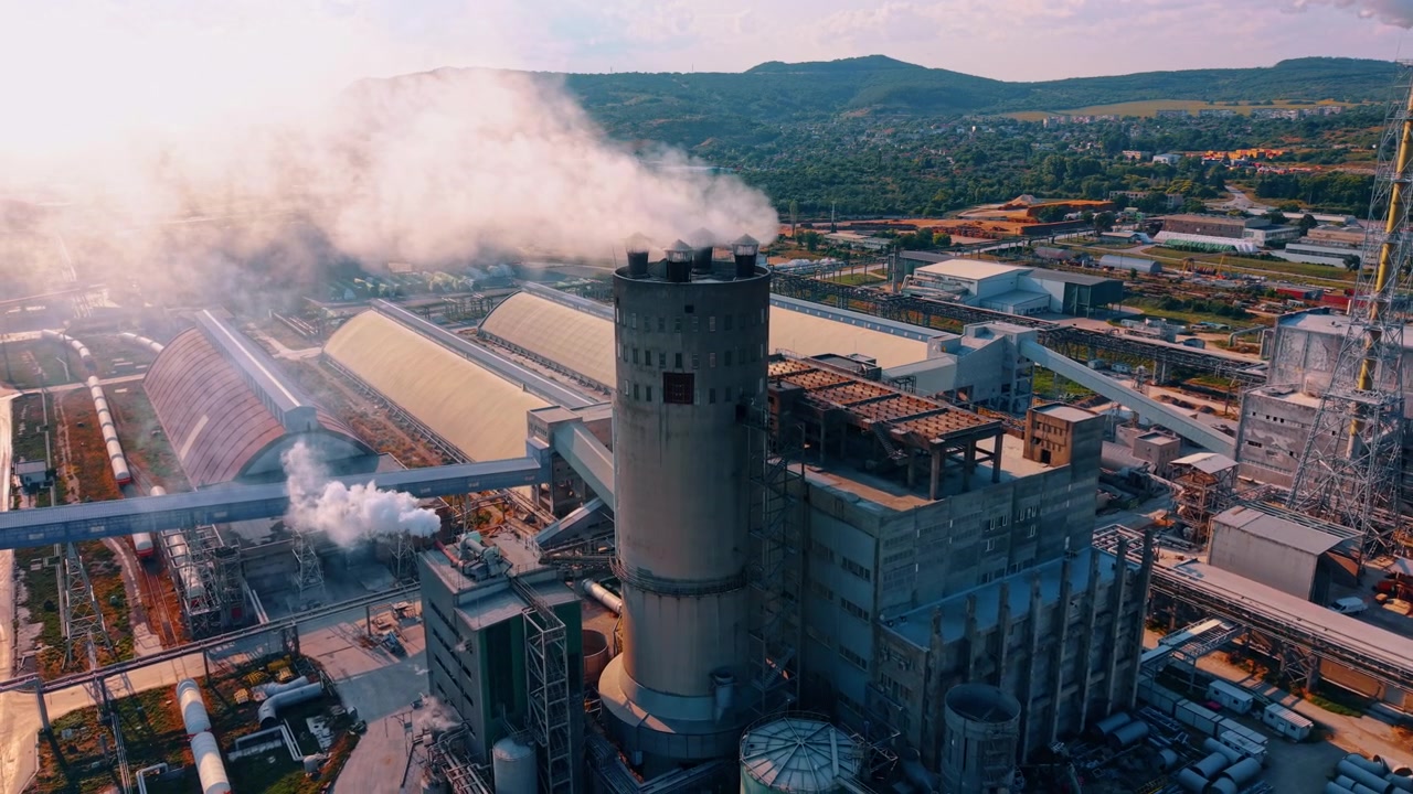 Torre industriale che rilascia fumo su un complesso di fabbriche. Una spessa colonna di fumo bianca si innalza da una ciminiera di cemento durante le operazioni diurne.
