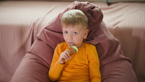 cute little boy relaxing on pink bean bag chair wearing yellow shirt eating ice cream cone while looking at camera with curious expression cozy indoor setting