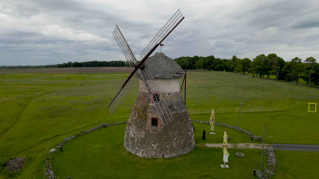 Aerial orbiting drone shot of a large and old stone windmill standing in a green grassy farm field during a cloudy day at Kuremaa. Horizon and treeline visible in the background and windmill flaps