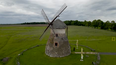 aerial orbiting drone shot of a large and old stone windmill standing in a green grassy farm field during a cloudy day at kuremaa. horizon and treeline visible in the background and windmill flaps