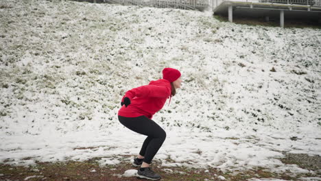 seitenansicht einer frau, die während des wintertrainings im freien auf schneebedecktem boden jump-squats durchführt, in einem roten hoodie und schwarzen handschuhen, umgeben von einem schneebeden hügel mit eisenbahn und einer nebligen atmosphäre