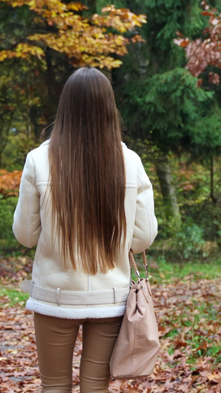 Stylish young lady with a big bag walks by the autumn park. Rear view. Woman turns around to the camera holding phone and paper cup in her hands. Vertical video