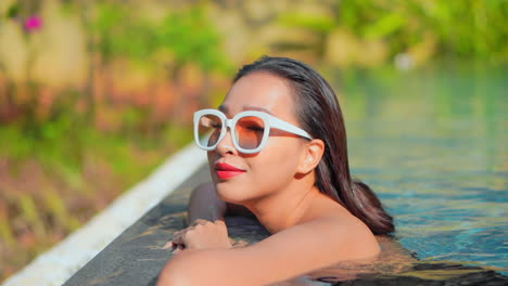 Vista-Lateral-De-Una-Turista-Con-Grandes-Gafas-De-Sol-Blancas,-Apoyada-En-El-Borde-De-Una-Piscina-Al-Aire-Libre-En-Algún-Destino-Tropical,-Poniendo-Su-Cabeza-En-El-Borde-Y-Sonriendo,-Luciendo-Feliz