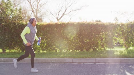 Video-Eines-älteren-Gemischtrassigen-Mannes-In-Sportkleidung-Und-Kopfhörern,-Der-Auf-Einer-Sonnigen-Straße-Läuft